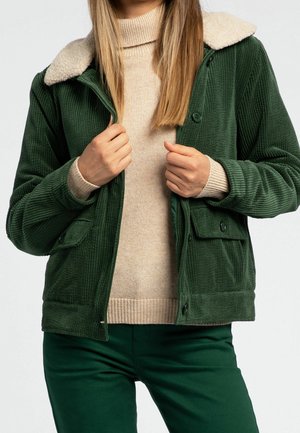 Femme portant une veste en velours côtelé verte avec un col sherpa crème par-dessus un pull à col roulé beige en maille et un pantalon vert.