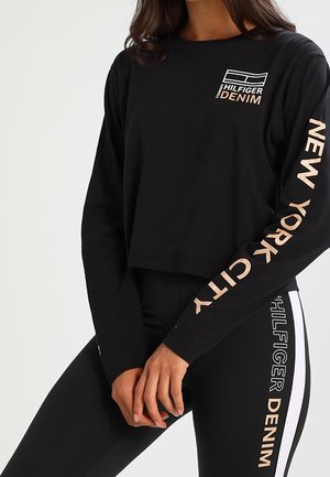 Kobieta nosząca czarny, długi top z napisem „New York City” na rękawie oraz dopasowane czarne legginsy z napisami „Hilfiger Denim”.