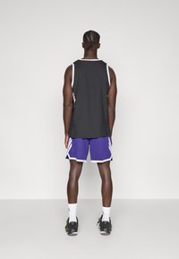 Μαύρο αμάνικο φανελάκι με λευκή λεπτομέρεια, συνδυασμένο με μωβ σορτς με λευκές πινελιές. Μαύρα αθλητικά παπούτσια και λευκές κάλτσες ολοκληρώνουν την εμφάνιση.