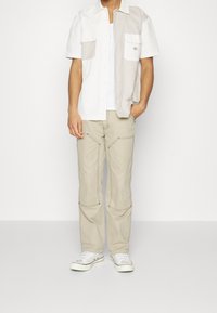 Camicia a maniche corte con bottoni bianca e grigio chiaro con due tasche sul petto, abbinata a pantaloni cargo beige con dettagli sulle cuciture e sneakers bianche.