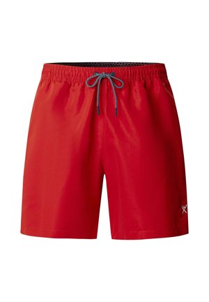 Hombre sin camisa de pie contra un fondo gris, vistiendo pantalones cortos de baño rojos con cordón gris y zapatos tipo slip-on negros.