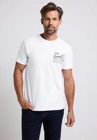 Bílá bavlněná tričko s krátkým rukávem, s designem levé kapsy na hrudi v černé barvě s grafickým textem a vlnkovým vzorem. Tmavě modrý denim.