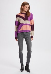 Maglione a blocchi di colore viola e pastello con vestibilità relaxed, maniche lunghe e polsini a coste, abbinato a jeans grigi e stivali neri con tacco.