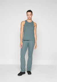 Ung kvinne står selvsikkert i en blå-grå treningstop og matchende tights med svarte joggesko mot en ensfarget bakgrunn.