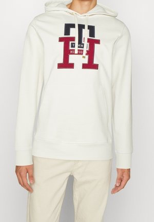 Biała bluza z kapturem z dużym czerwono-czarnym logo "TH" i napisem Tommy Hilfiger na piersi, noszona z jasnobeżowymi spodniami.