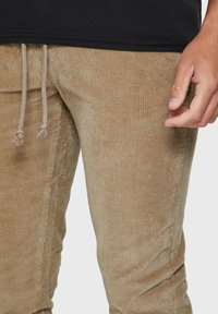 Beige corduroy broek met een geribbelde textuur, voorzien van een trekkoord taille en een strak aansluitend ontwerp. Een hand ligt op de stof.
