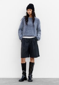 Pull en tricot bleu épais, bonnet noir, shorts noirs à rayures lâches, bottes montantes en cuir noir avec des boucles. Tenue décontractée.