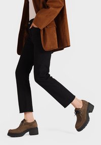 Scarpe platform in suede marrone con lacci e suole in gomma nera, abbinate a pantaloni neri a 3/4 e a una giacca in suede marrone. Design semplice.