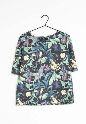 Chemise noire à manches courtes avec des motifs floraux et de feuilles verts, bleus, violets et crème, suspendue sur un cintre en fil noir sur fond blanc.