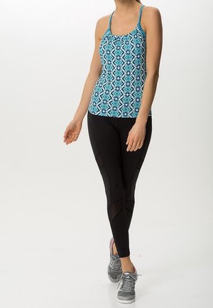 Femme portant un débardeur bleu à motifs, des leggings noirs avec des détails en mesh et des baskets grises, debout sur un fond blanc.