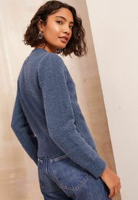 Mīksts, zils krekls ar garām piedurknēm un pieguļošu dizainu, valkāts ar augstiem džinsiem. Strukturēta auduma un apaļa dekoltē.