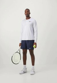 Herre treningsantrekk som består av en langermet hvit topp med knapp, marineblå shorts og hvite joggesko, mens han holder en tenniskølle og ball.