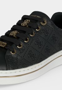 Baskets noires avec motif de logo embossé, lacets noirs ornés de charms décoratifs dorés, et semelle blanche avec une bande dorée.