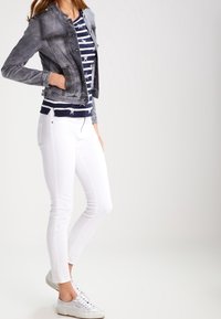 Donna con una giacca di jeans nera sbiadita indossata sopra una camicia a righe blu con motivo a stelle, jeans skinny bianchi e sneaker bianche, in piedi con una mano in tasca.