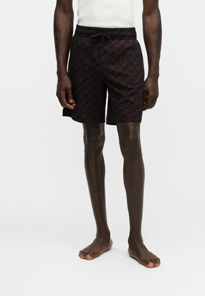 Homme portant un short noir avec le texte rouge répété "HUGO", associé à un haut blanc sans manches, debout pieds nus devant un fond uni.