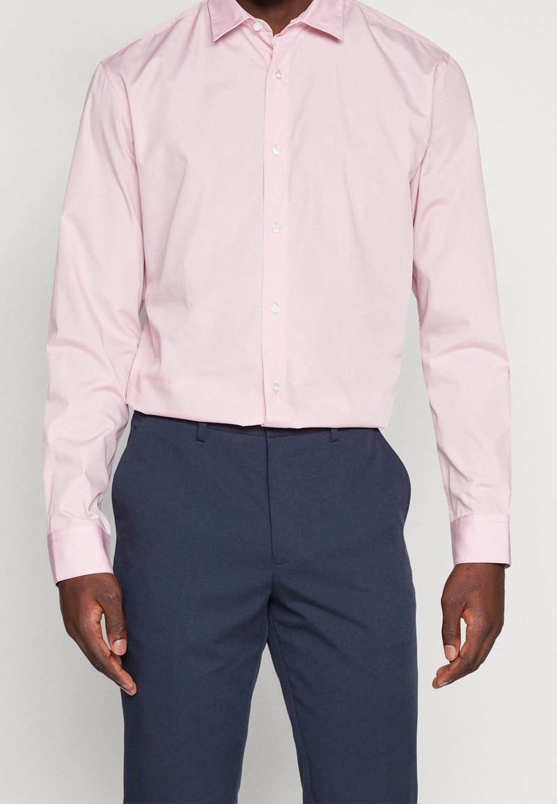 Homme portant une chemise rose pâle à manches longues boutonnée, rentrée dans un pantalon bleu marine foncé, debout devant un fond uni.
