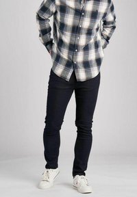 Mężczyzna w koszuli w kratę z guzikami, ciemnych dżinsach i białych trampkach, stojący z rękami w kieszeniach na neutralnym tle.