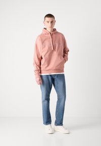 Rosa Kapuzenpullover mit einer Vordertasche und Kordelzug-Haube, mit erhabener "HUGO"-Aufschrift. Kombiniert mit blauen Jeans und weißen Sneakern.