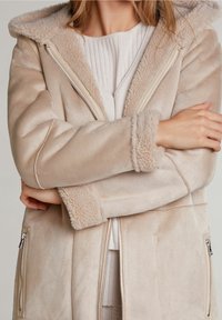 Kvinde iført en beige pelsjakke med lynlåslommer og foldede arme, der viser bløde teksturerede manchetter og hættefôr.