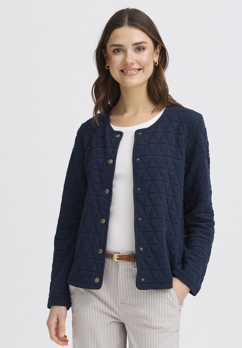 Jeune femme portant une veste matelassée bleu marine sur une chemise blanche, un pantalon rayé et une ceinture tressée marron, souriante avec une main dans la poche.