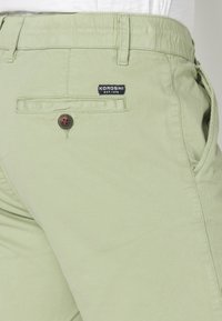 Pantalones cortos de algodón verde claro con cremallera, bolsillos laterales y un bolsillo trasero que presenta un botón oscuro y una etiqueta con el logo.