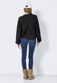 Zwarte gebreide trui met gevallen schouders, geribbelde boorden, gecombineerd met blauwe skinny jeans en tan kleurige gewatteerde enkellaarzen. Witte baseballcap achterstevoren gedragen.