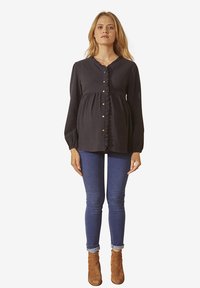Schwangere Frau in einer schwarzen, gerüschten Bluse mit Knöpfen, blauen Skinny-Jeans und braunen Stiefeletten, die vor einem einfarbigen Hintergrund steht.