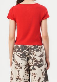 Top corto rojo de manga corta con un escote redondeado, combinado con pantalones beige con un patrón de manchas marrones y dos bolsillos traseros.