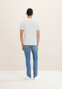 Hvid kortærmet t-shirt parret med medium vaskede jeans og hvide sneakers, set fra ryggen mod en lys beige baggrund.