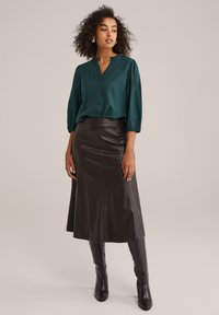 Grüne Bluse mit Knopfleiste und 3/4-Ärmeln sowie einem gerafften Ausschnitt, kombiniert mit einem schokoladenbraunen Leder-Midi-Rock und hohen braunen Stiefeln.