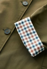 Manche de veste vert olive avec boutons visibles et revers replié présentant un motif à carreaux bleu, marron et blanc.