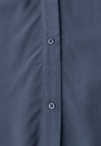 Gros plan sur le devant d'une chemise bleue montrant deux petits boutons ronds fixés dans les boutonnières. Le tissu est lisse avec une texture subtile.