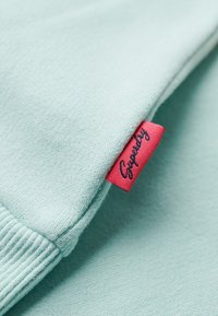 Tissu bleu clair avec un bord-côte et une petite étiquette rouge portant la mention "Superdry" cousue dans la couture.