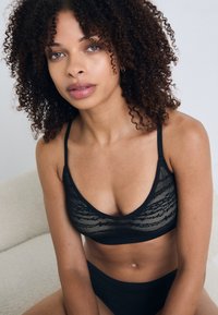Svart blonderbralette med justerbare stropper og et mykt, sømløst design. Kombinert med matchende svarte truser, plassert på en nøytral bakgrunn.