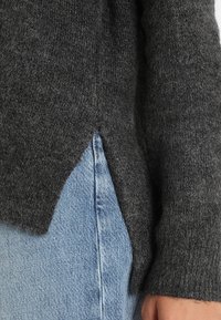 Pull gris en maille avec une texture douce et une fente latérale, associé à un jean en denim bleu clair. Mise en avant des détails du matériau et du design.
