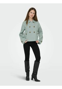 Jeune femme portant une veste vert clair à double boutonnage, un pantalon noir slim et des bottes noires à talons montant jusqu'aux genoux, debout les mains près des poches.