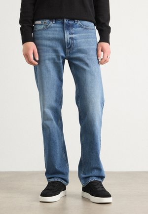 Homme portant un jean droit bleu avec des baskets noires et un pull noir, debout devant un fond clair uni.