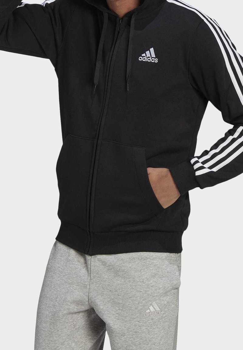 Sweat à capuche zippé noir avec un design à trois bandes blanches sur les manches, des poches avant et un logo brodé. Associé à un pantalon de jogging gris.