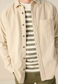 Beige corduroy overhemd met knopen en een kraag, voorzien van een borstzak en bruine knopen, gedragen over een groen-wit gestreept sweater.