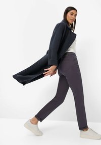 Mørkegrå frakke med høj krave, båret over en hvid top og mørkelilla bukser. Lyse sneakers fuldender outfittet.