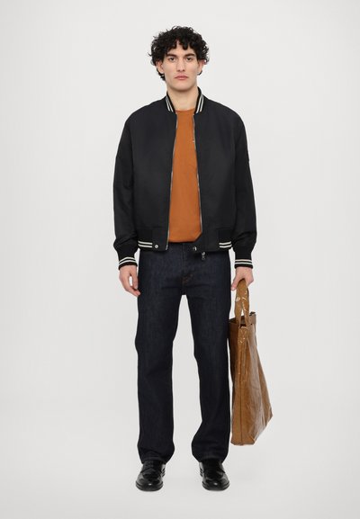 Giubbotto bomber nero con strisce bianche, t-shirt arancione, jeans scuri e scarpe nere. Porta una borsa di carta marrone.