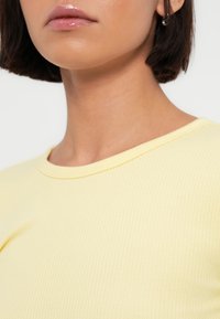 Κίτρινο κορμάκι με ραβδώσεις, που διαθέτει στρογγυλή λαιμόκοψη. Το ύφασμα φαίνεται μαλακό με λεπτές κάθετες γραμμές. Φαίνεται το σκούρο μαλλί του ατόμου.