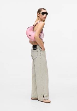 Femeie purtând ochelari de soare oversized, un top roz cu imprimeu floral, blugi largi de culoare deschisă și având pe umăr o geantă roz în formă de semilună.