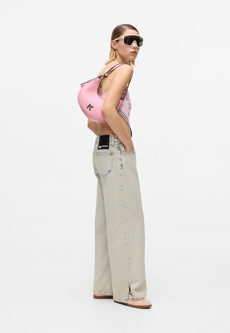 Γυναίκα που φοράει πολύ μεγάλα γυαλιά ηλίου, ροζ τοπ με λουλούδια, φαρδιά ανοιχτόχρωμα τζιν και κρατάει ροζ τσάντα σε σχήμα ημισελήνου στον ώμο της.