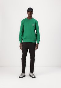 Felpa verde con logo, pantaloni neri e sneakers grigie. La felpa ha un collo a giro e polsini a coste. Il cappellino è nero.