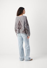 Gestrickter Pullover in Grau mit einem strukturierten Muster in Dunkelbraun. Oversized-Schnitt, gerippter Saum und dégradé Schultern. Kombiniert mit hellblauen Jeans.