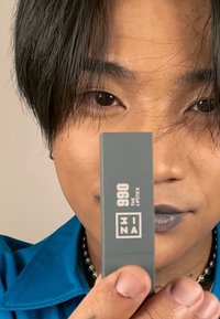 Grå leppestift tube merket "990 THE LIPSTICK" med matte finish. Bakgrunnen er nøytral, og subjektet har på seg en blå skjorte og et perlekjede.