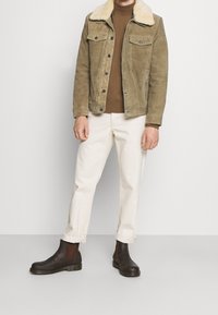 Hnědá semišová bunda s falešným límcem z beránka, v kombinaci s hnědým svetrem a krémovými kalhotami. Modelka má na nohou hnědé chelsea boty.