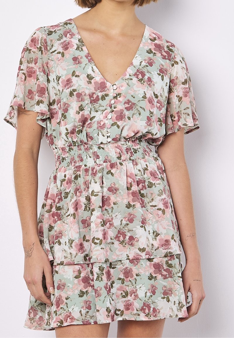 Femme portant une robe courte fleurie avec des fleurs roses et blanches, manches volantes, col en V, boutonnage devant et taille élastique.