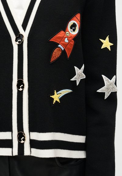 Μαύρο ζακέτα με λευκές ρίγες, διακοσμημένη με πολύχρωμα κεντητά μπαλώματα πυραύλου, αστεριών και ενός γελαστού προσώπου. Τα κουμπιά είναι διακοσμητικά.